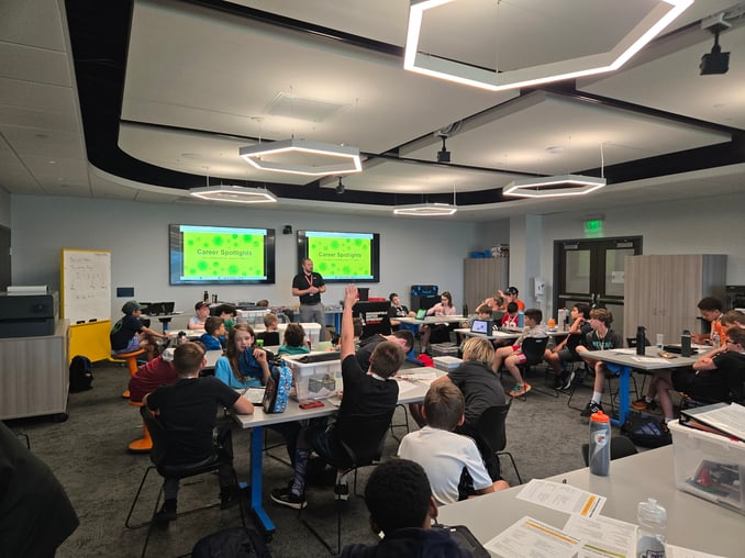 About 25 middle-grade students sit in a classroom on orange stools or black chairs and at white tabl…
