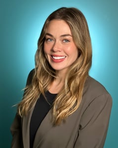 A headshot of a woman with light brown hair who is smiling and wearing a tan jacket with black shirt.